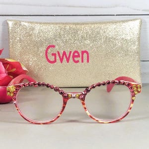 May include: Eyeglasses with a pink and orange patterned frame embellished with small red and yellow rhinestones along the top rim. Behind the glasses sits a rectangular gold glitter pouch with the name Gwen printed in pink lettering.
