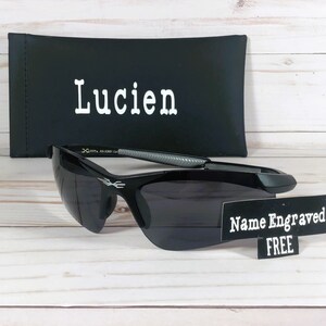 May include: Black sunglasses with a gray and silver frame. The sunglasses are on a wooden surface with a black case that says "Lucien" on it. A sign says "Name Engraved FREE".