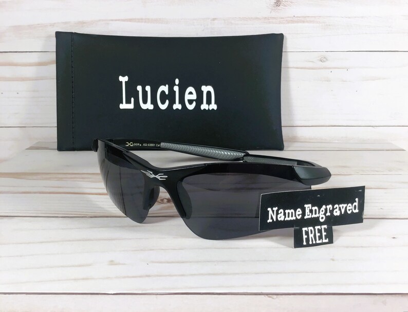 May include: Black sunglasses with a gray and silver frame. The sunglasses are on a wooden surface with a black case that says "Lucien" on it. A sign says "Name Engraved FREE".