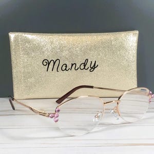 May include: Gold-rimmed spectacles with clear lenses and pink embellishments, placed before a gold, textured case. The case is embroidered with the name "Mandy" in black script. The glasses have a delicate, feminine design.