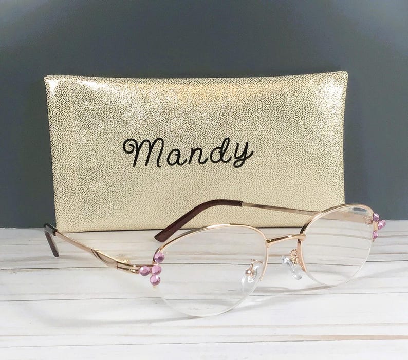 May include: Gold-rimmed spectacles with clear lenses and pink embellishments, placed before a gold, textured case. The case is embroidered with the name "Mandy" in black script. The glasses have a delicate, feminine design.