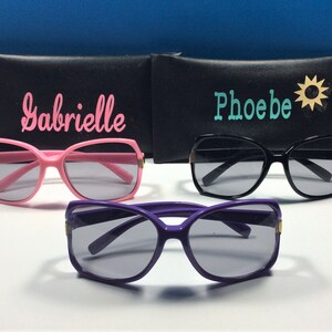 May include: Three pairs of sunglasses with black cases. The cases have the names "Gabrielle", "Phoebe", and a sun icon on them. The sunglasses are pink, black, and purple.