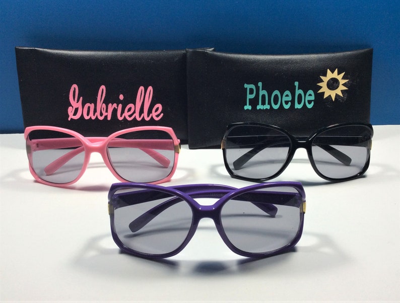 May include: Three pairs of sunglasses with black cases. The cases have the names "Gabrielle", "Phoebe", and a sun icon on them. The sunglasses are pink, black, and purple.
