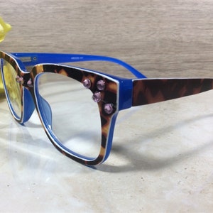 May include: Eyeglasses with a blue frame and temples, featuring a tortoiseshell pattern on the front and temples. The frames are adorned with pink rhinestones. The lenses are clear, and the glasses are set against a neutral background.