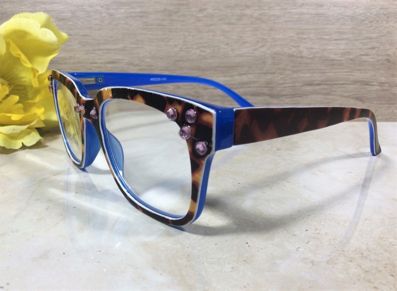 May include: Eyeglasses with a blue frame and temples, featuring a tortoiseshell pattern on the front and temples. The frames are adorned with pink rhinestones. The lenses are clear, and the glasses are set against a neutral background.