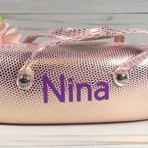 May include: A pink, textured cosmetic bag with a handle and the name "Nina" in purple lettering. The bag has silver-coloured metal accents and a matching handle. The background includes pink flowers and a white wooden surface.