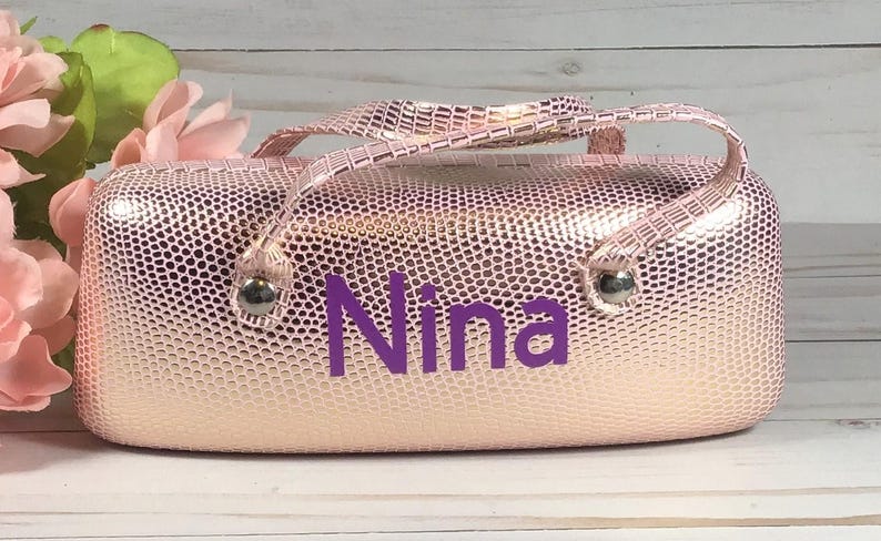 May include: A pink, textured cosmetic bag with a handle and the name "Nina" in purple lettering. The bag has silver-coloured metal accents and a matching handle. The background includes pink flowers and a white wooden surface.