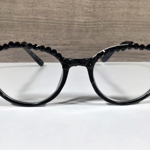 Puede incluir: Gafas negras con un dise&ntilde;o de montura redondeada y una hilera de peque&ntilde;as cuentas o pedrer&iacute;a negra circular incrustada a lo largo del borde superior de los aros. Las gafas tienen lentes transparentes y patillas negras finas en los laterales.