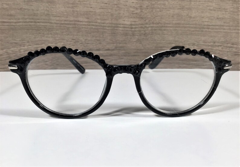 Puede incluir: Gafas negras con un dise&ntilde;o de montura redondeada y una hilera de peque&ntilde;as cuentas o pedrer&iacute;a negra circular incrustada a lo largo del borde superior de los aros. Las gafas tienen lentes transparentes y patillas negras finas en los laterales.