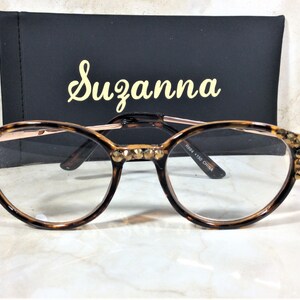 May include: Round eyeglasses with a tortoiseshell pattern frame and gold-colored accents. The frames are embellished with small, sparkling crystals. The glasses are set in front of a black case with the name "Suzanna" in gold script.