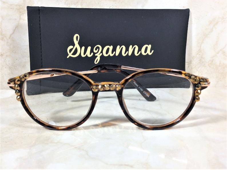 May include: Round eyeglasses with a tortoiseshell pattern frame and gold-colored accents. The frames are embellished with small, sparkling crystals. The glasses are set in front of a black case with the name "Suzanna" in gold script.