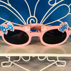 May include: Pink sunglasses with black lenses, decorated with blue flower and butterfly accents. The frames feature heart and bow details. A fun accessory for children.