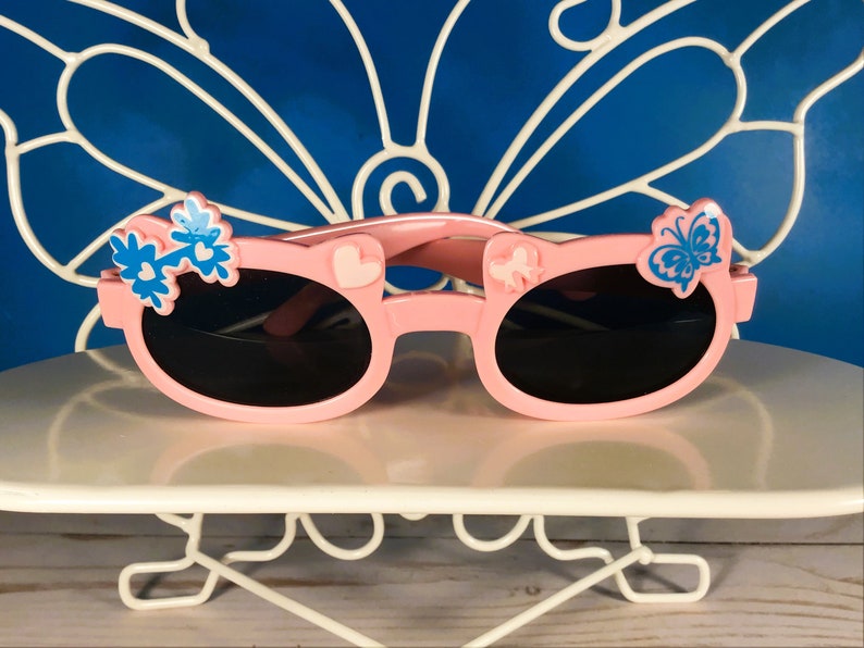 May include: Pink sunglasses with black lenses, decorated with blue flower and butterfly accents. The frames feature heart and bow details. A fun accessory for children.