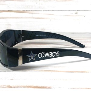 May include: Black sunglasses with the word "COWBOYS" in white lettering and a silver star logo on the arm. The sunglasses are made in China.