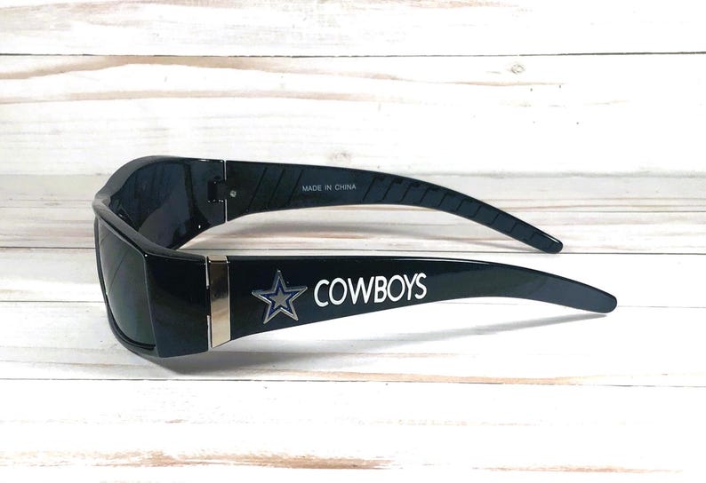 May include: Black sunglasses with the word "COWBOYS" in white lettering and a silver star logo on the arm. The sunglasses are made in China.