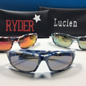 May include: Three pairs of sunglasses with camouflage patterned frames. The sunglasses have different coloured lenses: red, blue and green. The sunglasses are displayed on top of two black cases with the names "Ryder" and "Lucien" printed on them.