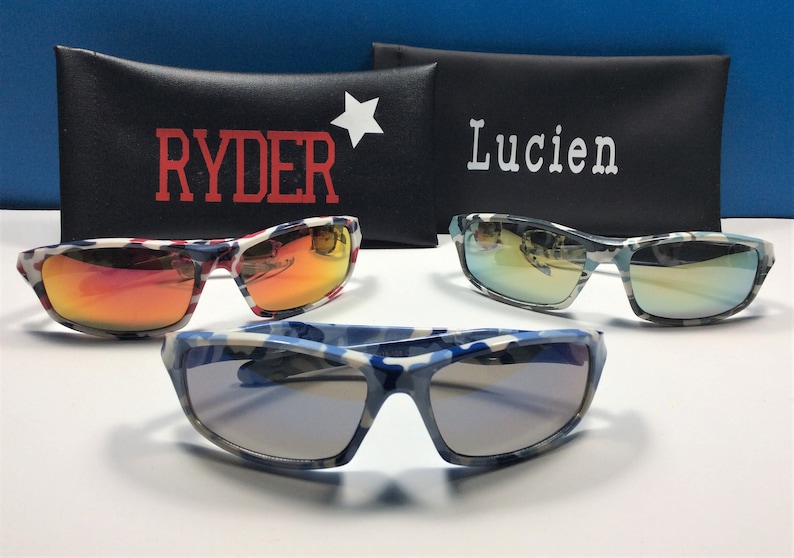 May include: Three pairs of sunglasses with camouflage patterned frames. The sunglasses have different coloured lenses: red, blue and green. The sunglasses are displayed on top of two black cases with the names "Ryder" and "Lucien" printed on them.