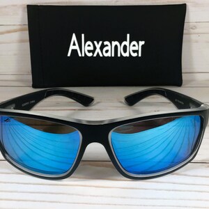 K&ouml;nnte beinhalten: Schwarze Sonnenbrille mit blauen verspiegelten Gl&auml;sern und silbernem Rahmen. Die Sonnenbrille liegt auf einer wei&szlig;en Holzoberfl&auml;che. Der Markenname "Alexander" ist auf einem schwarzen Etui aufgedruckt.
