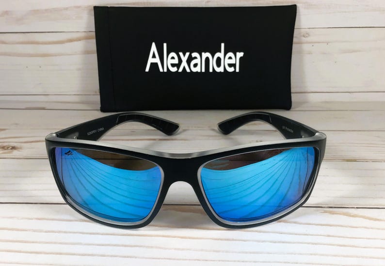 K&ouml;nnte beinhalten: Schwarze Sonnenbrille mit blauen verspiegelten Gl&auml;sern und silbernem Rahmen. Die Sonnenbrille liegt auf einer wei&szlig;en Holzoberfl&auml;che. Der Markenname "Alexander" ist auf einem schwarzen Etui aufgedruckt.