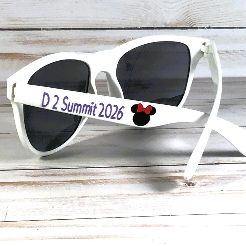 May include: White sunglasses with dark lenses. The left arm of the sunglasses has the text "D 2 Summit 2026" in purple, and a black Minnie Mouse head with a red bow. The sunglasses are on a light wood surface.