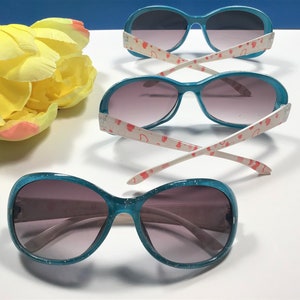 May include: Three pairs of turquoise blue sunglasses with glitter on the frames. The sunglasses have pink heart patterns on the white arms.