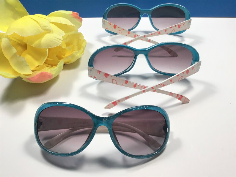 May include: Three pairs of turquoise blue sunglasses with glitter on the frames. The sunglasses have pink heart patterns on the white arms.
