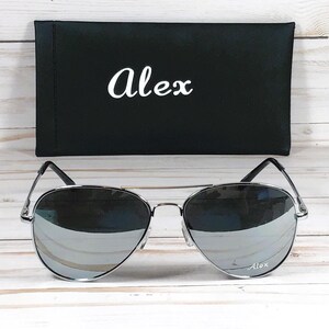 May include: Silver aviator sunglasses with mirrored lenses, resting in front of a black case. The case has the name "Alex" in white script. The sunglasses have a silver frame and the name "Alex" is also on the lens.