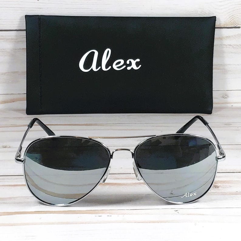 May include: Silver aviator sunglasses with mirrored lenses, resting in front of a black case. The case has the name "Alex" in white script. The sunglasses have a silver frame and the name "Alex" is also on the lens.