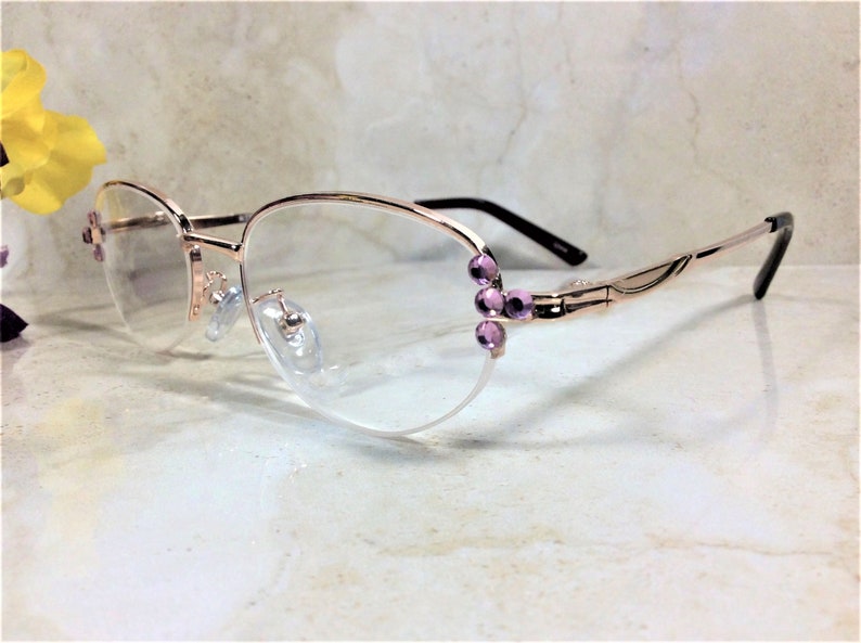 Pu&ograve; includere: Occhiali in oro rosa con una montatura delicata e lenti trasparenti. Le aste sono nere e la montatura &egrave; adornata da piccoli accenti viola a forma di gioiello. Gli occhiali hanno una forma ovale classica.
