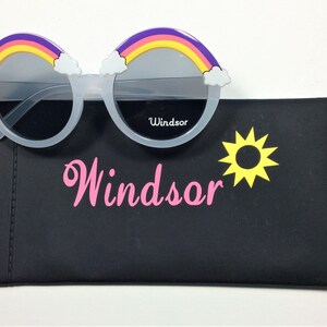 Puede incluir: Gafas de sol blancas con un dise&ntilde;o de arco&iacute;ris y nubes en el marco. Las gafas de sol vienen con una funda de transporte negra con un sol amarillo y la palabra "Windsor" impresa en rosa.