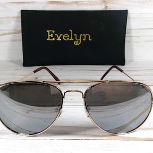 May include: Gold aviator sunglasses with mirrored lenses. The sunglasses are on a white wooden background.