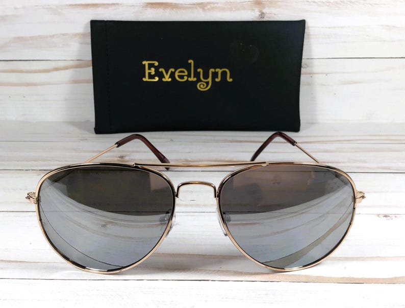 May include: Gold aviator sunglasses with mirrored lenses. The sunglasses are on a white wooden background.