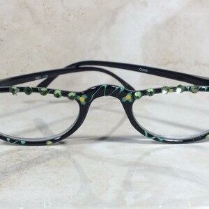 May include: Black half-frame spectacles with clear lenses. The frames feature green and yellow floral patterns and green rhinestones along the top. The arms are black and the text "China" is visible on the arm.