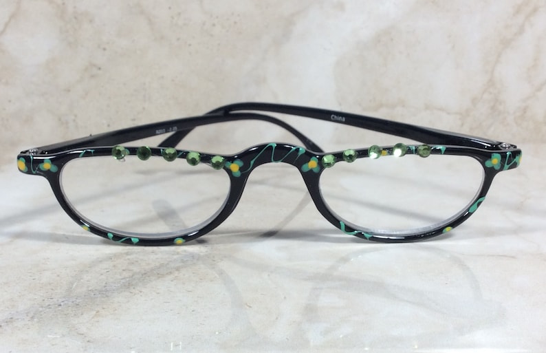 May include: Black half-frame spectacles with clear lenses. The frames feature green and yellow floral patterns and green rhinestones along the top. The arms are black and the text "China" is visible on the arm.