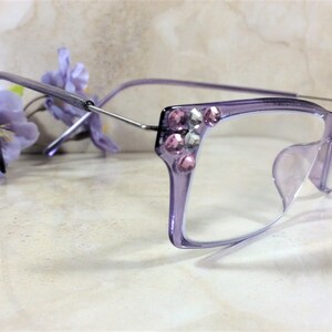 May include: Light purple translucent eyeglasses featuring a rectangular frame design. The outer corners of the frames are embellished with a cluster of small, round, pink and clear faceted rhinestones. The temples are made of thin silver metal.