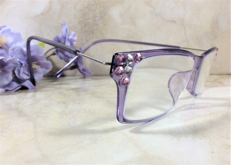 May include: Light purple translucent eyeglasses featuring a rectangular frame design. The outer corners of the frames are embellished with a cluster of small, round, pink and clear faceted rhinestones. The temples are made of thin silver metal.