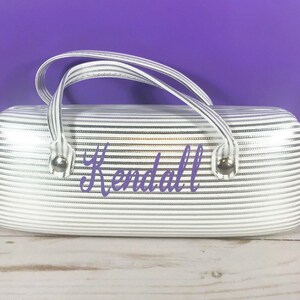 May include: A white and silver striped glasses case with a handle. The case has the name "Kendall" in purple script. The case has silver metal accents and a white handle.