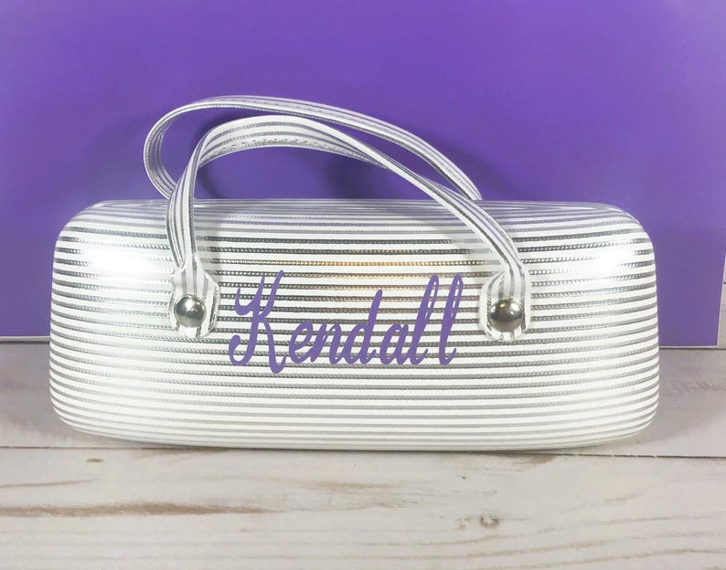 May include: A white and silver striped glasses case with a handle. The case has the name "Kendall" in purple script. The case has silver metal accents and a white handle.