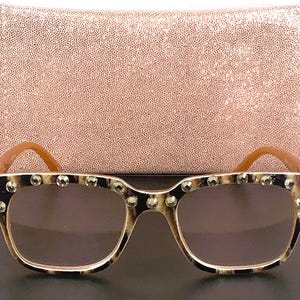 Puede incluir: Gafas de sol cuadradas con montura de carey y tachuelas plateadas en la parte superior. Las lentes son marrones. Las patillas son de color marr&oacute;n claro. En el fondo, un bolso de mano rosa texturizado.