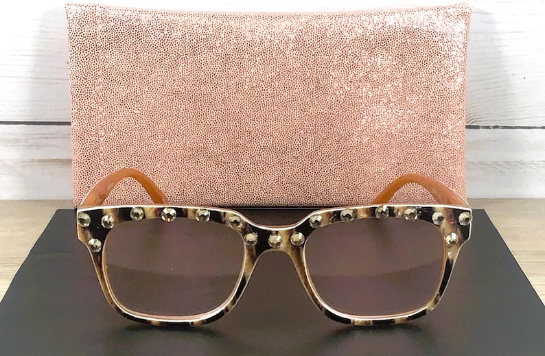Puede incluir: Gafas de sol cuadradas con montura de carey y tachuelas plateadas en la parte superior. Las lentes son marrones. Las patillas son de color marr&oacute;n claro. En el fondo, un bolso de mano rosa texturizado.