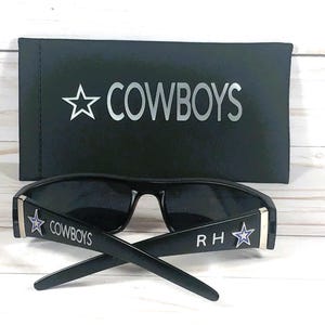 May include: Black sunglasses with the word "COWBOYS" and a star logo on the temples. The sunglasses are paired with a black case that also features the word "COWBOYS" and a star logo.