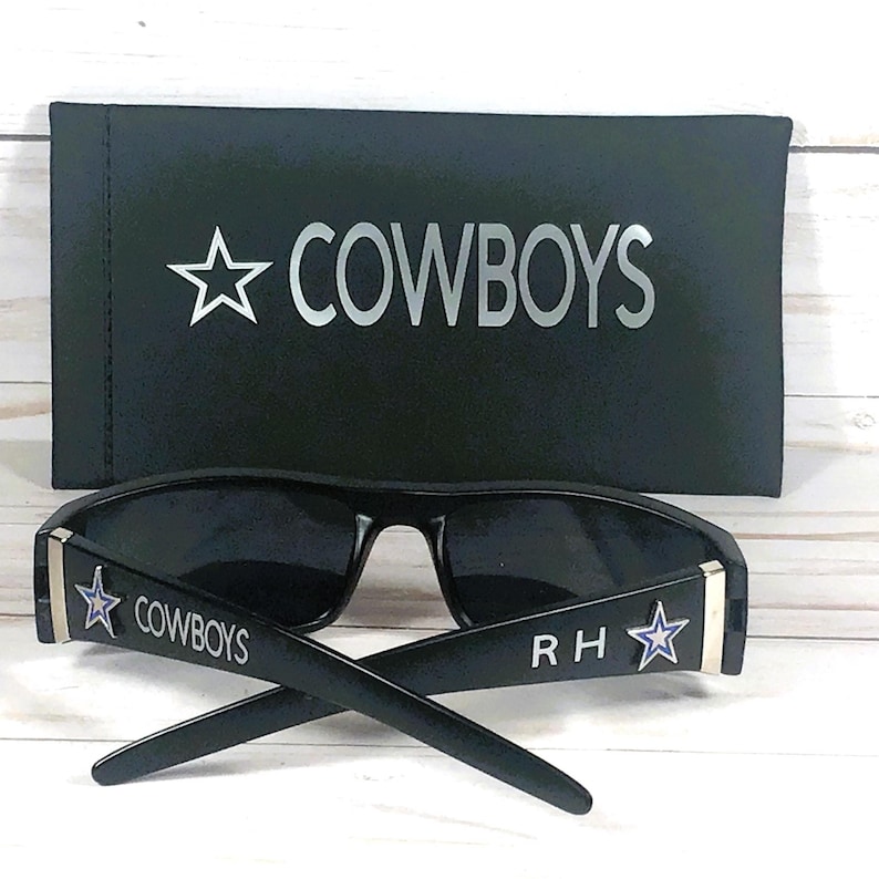 May include: Black sunglasses with the word "COWBOYS" and a star logo on the temples. The sunglasses are paired with a black case that also features the word "COWBOYS" and a star logo.