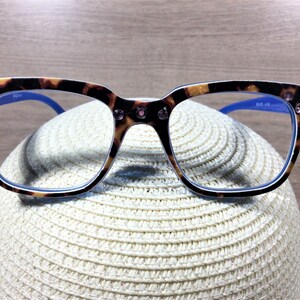 May include: Eyeglasses with a tortoiseshell pattern frame, adorned with pink rhinestones at the corners. The frames have blue arms and are resting on a white straw hat. The glasses have a square shape.