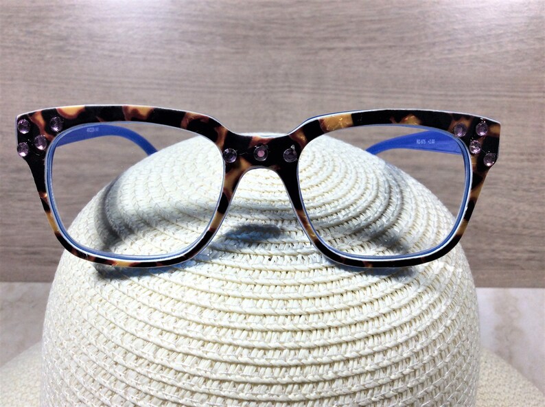 May include: Eyeglasses with a tortoiseshell pattern frame, adorned with pink rhinestones at the corners. The frames have blue arms and are resting on a white straw hat. The glasses have a square shape.