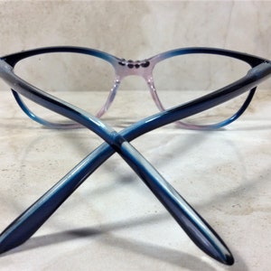 Puede incluir: Gafas con forma de ojo de gato, con un degradado de colores. Las monturas transicionan de negro a azul y luego a un rosa suave en el puente nasal. Las patillas de las gafas est&aacute;n cruzadas, mostrando el degradado azul.