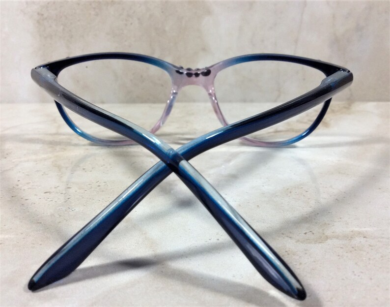 Puede incluir: Gafas con forma de ojo de gato, con un degradado de colores. Las monturas transicionan de negro a azul y luego a un rosa suave en el puente nasal. Las patillas de las gafas est&aacute;n cruzadas, mostrando el degradado azul.