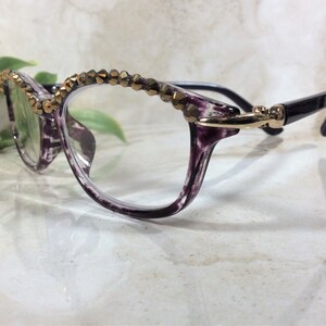 May include: Spectacles featuring a translucent purple and black marbled frame with a row of metallic bronze-toned faceted beads along the top rim. The temples are solid black and connect to the frame with a gold-toned metal hinge detail.