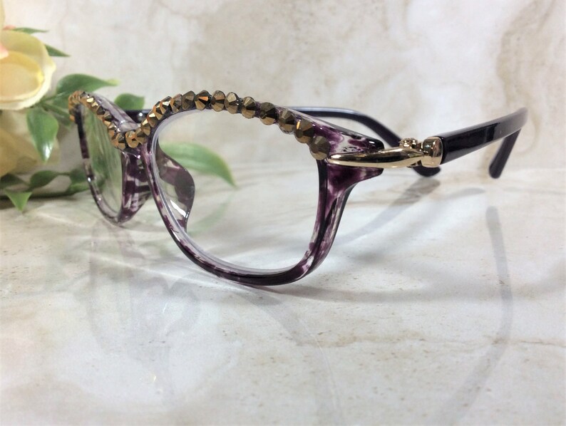 May include: Spectacles featuring a translucent purple and black marbled frame with a row of metallic bronze-toned faceted beads along the top rim. The temples are solid black and connect to the frame with a gold-toned metal hinge detail.