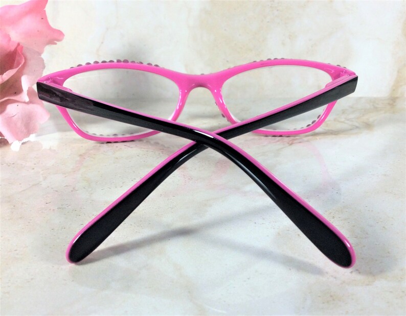 Pu&ograve; includere: Occhiali rosa acceso con aste nere e lenti trasparenti. Le montature sono decorate con piccoli abbellimenti scintillanti. Gli occhiali hanno un design moderno a occhi di gatto.