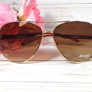 May include: Gold-framed aviator sunglasses with brown gradient lenses. The sunglasses have a double bridge and decorative accents on the temples. The text "Italian Design" and "Alcocer" are visible on the frame.